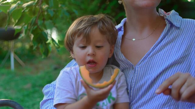 Toddler eating cantaloupe on mother&rsquo;s lap outdoors in the garden, intimate family bonding reflecting comfort, maternal care and resilience during cancer