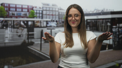 Fototapeta premium Woman shrugging with palms up and bare hands beside canal boats on a city street, smiling with closed eyes; playful ease.