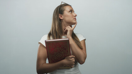 Woman holding ornate red hardcover book, finger pressed to chin in studio with glasses on head and...
