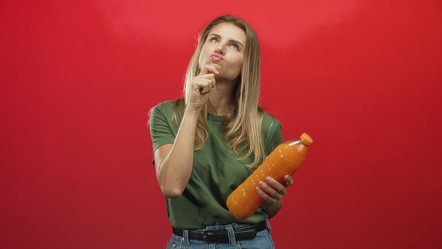 Woman holding large orange juice bottle with puckered lips and pinching fingers while examining cap in studio; taste decision curiosity.