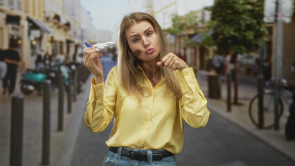Fototapeta premium Woman holding a toy airplane in her right hand, left fist by chin, pouting expression on a busy street; travel frustration.