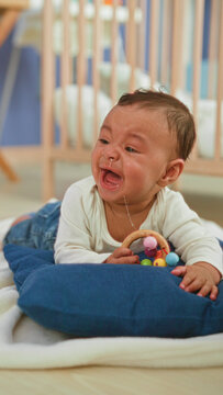 Hispanic baby boy holding wooden rattle, crying with tears and drool while caregiver wipes his face in building; distress care.