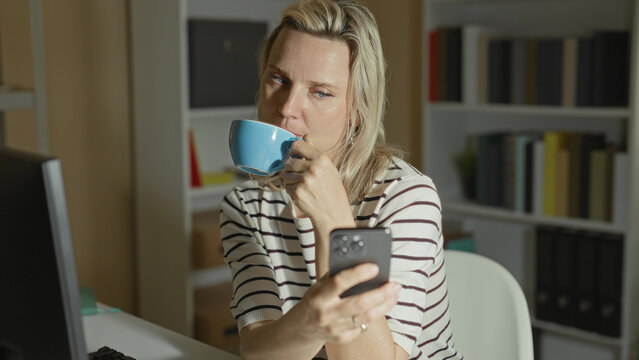 Woman holding smartphone and sipping from a blue mug while seated at a computer in an office building; late night contemplation.