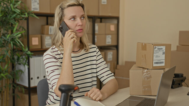 Woman holding phone to ear and pen beside stacked parcels and laptop in packing area building; small business stress.
