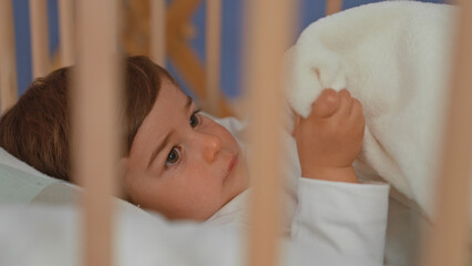 Boy in wooden crib lies on back reaching hand to soft white blanket while gazing upward inside a building  calm comfort. © Krakenimages.com