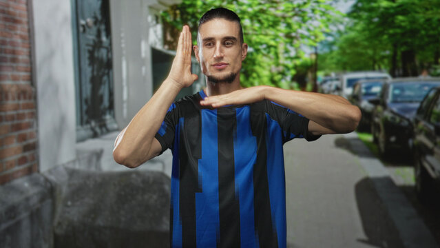 Man in blue and black soccer jersey forms timeout hand gesture with visible hands and forearms on a leafy street with parked cars and brick building; team pride confidence.
