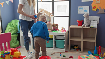 Fototapeta premium Boy holds blue ball and reaches with hand while woman stands nearby in a playroom building; playful learning.