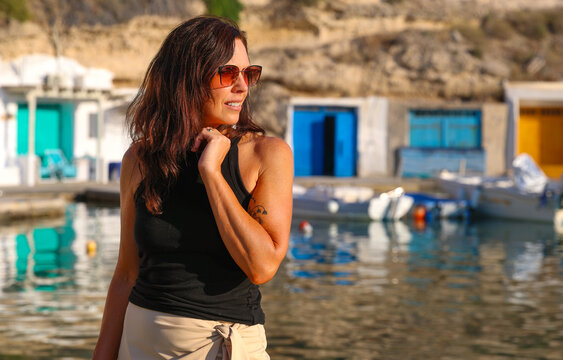 Woman standing beside the waterfront in Mandrakia, Milos, Greece, with colorful boat garages, calm harbor waters, and traditional Cycladic island architecture.