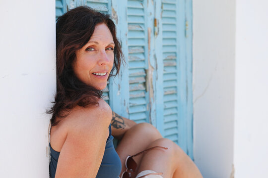 Woman seated beside weathered blue shutters in Plaka,, Milos, Greece, showcasing traditional Cycladic architecture and relaxed island lifestyle.
