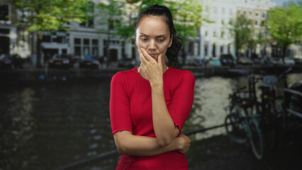 Fototapeta premium Woman in red top touching her face with hand, arms crossed, standing by parked bicycle along canal street with buildings and trees behind; quiet contemplation.