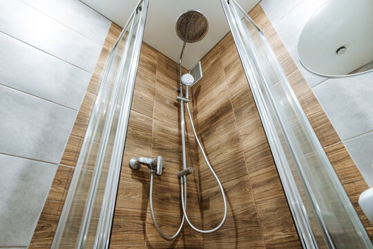 Chrome showerhead and handheld unit in tiled bathroom corner with glass enclosure, wood-effect walls, gray accents, reflective metal fixtures and clean modern interior detail