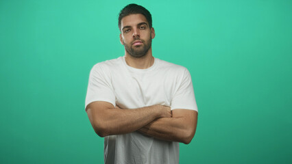 Fototapeta premium Hispanic man with beard stands with arms crossed, bare forearms visible, wearing white t shirt against a green studio backdrop, neutral serious expression; confidence solitude resolve.