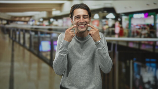 Man points index fingers to cheeks smiling in a busy mall building with metal railing, shops and blurred shoppers behind him, casual grey crewneck and jewelry; playful joy.