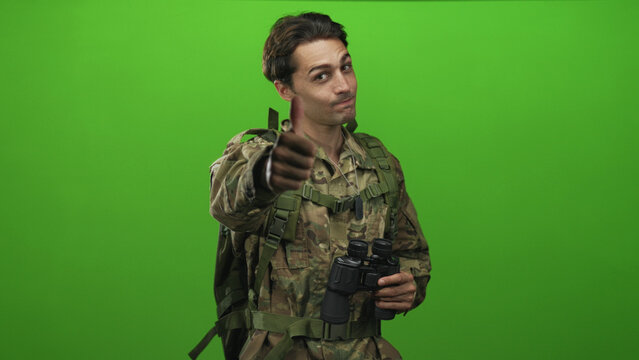 Soldier in camouflage uniform wearing backpack and dog tags, holding binoculars and giving thumbs up to camera in studio; approval duty morale.