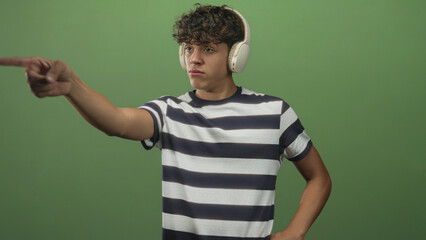 Fototapeta premium Teenage man wearing headphones points finger toward camera in a green studio; concentration intent focus.
