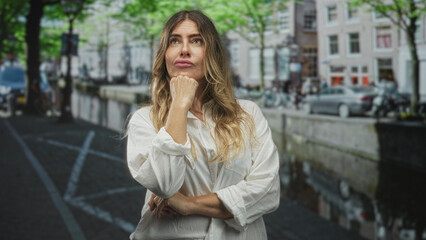 Fototapeta premium Woman with hand under chin, eyes closed and thinking while standing on a street by a canal and parked cars; contemplation.