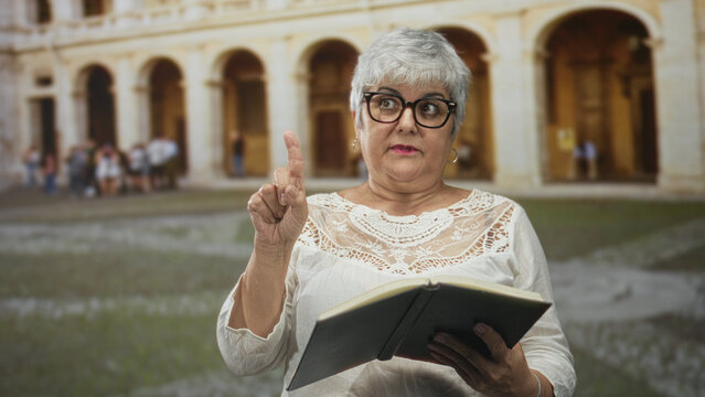 Woman reader wearing glasses with grey hair holding book, hand to chest and pointing finger, mouth open in a historic building courtyard; dramatic surprise.