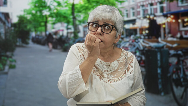 Senior woman reader with grey hair holds open book and bites fingernail with left hand on a city street lined with bicycles and cafes; anxiety.