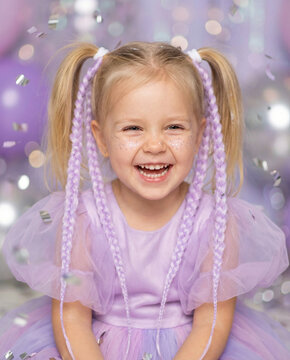 Happy child girl wearing purple hair braids with glitter on face. Celebration concept for birthday party. Fashionable hairstyle for kid with colorful accessories and confetti background.
