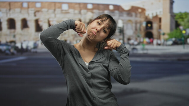 Young woman pinches her cheek and pulls down her mouth with fingers in front of an ancient building on a busy street; self mockery frustration.