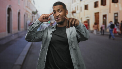 Fototapeta premium Black man pointing with index finger, touching temple in one frame, smiling while walking through a crowded historic street with blurred pedestrians; confidence connection.