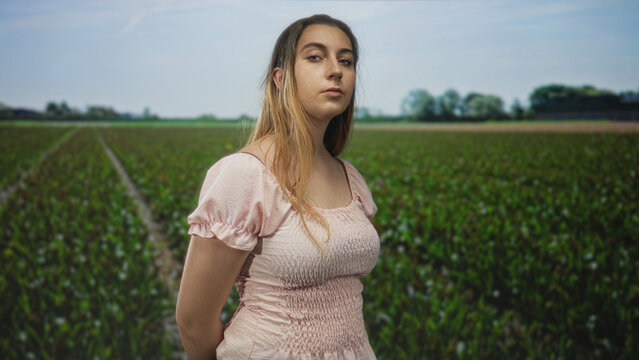 Woman wearing a pink smocked dress stands with hands behind her back in a crop field under a blue sky; quiet confidence.
