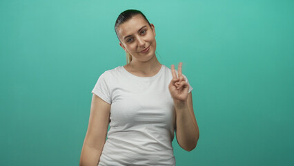 Fototapeta premium Woman in white tee smiles with tilted head and holds up a two finger peace sign with right hand against a teal studio backdrop; playful confidence.