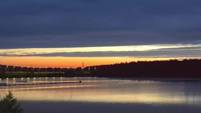Calm river with reflecting orange sunset, dark silhouettes of trees and distant power lines against twilight sky, rows of lit streetlights along the embankment, tranquil evening waterscape