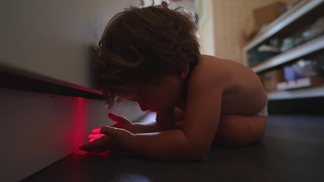 Curious toddler crouches on the floor and reaches into a red glow beneath a cabinet at home, exploring light, color and shadow through focused sensory discovery