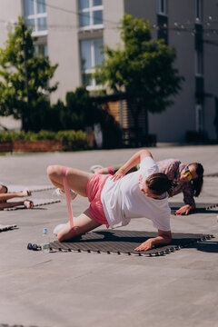 Woman performing side plank exercise with a resistance band during an outdoor fitness class in an urban environment. Healthy lifestyle, strength training, functional workout, and active recreation.