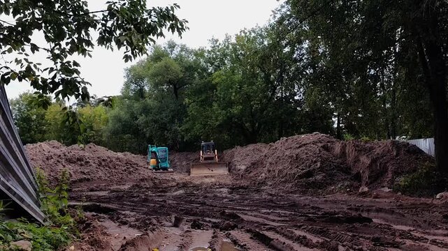 Wide view of large sand piles at municipal construction site surrounded by forest with two tractors in the background, infrastructure development,heavy machinery, earthmoving operations, summer season