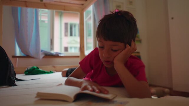 Focused child traces words in an open story book while resting on a bed at home, strengthening reading skills, comprehension, curiosity and independent education