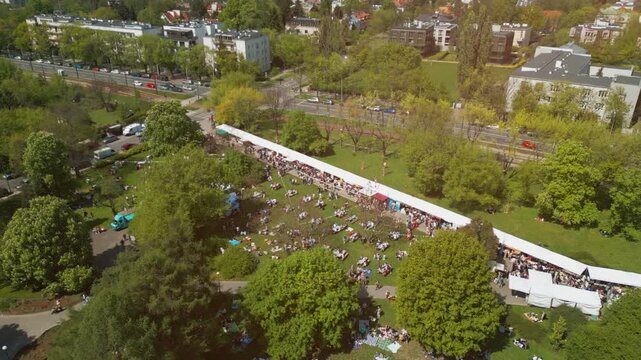 city street food festival event at green park at summer, tents of food market at green meadow, crowd walking and sitting at picnic, aerial view 4k