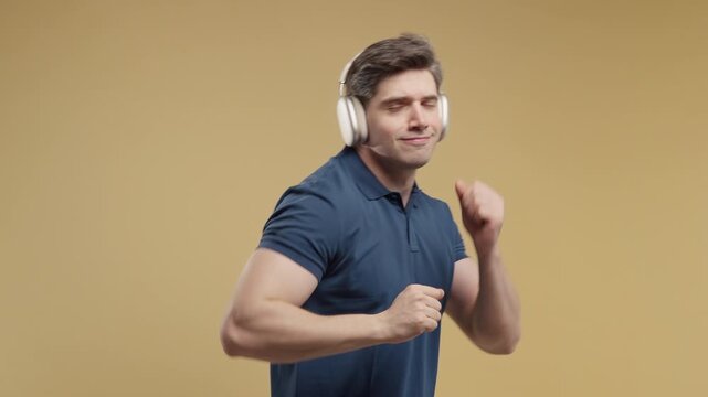 Happy adult man enjoying music through wireless headphones while dancing. Modern technology, wellness and joyful lifestyle. Studio portrait on a beige background