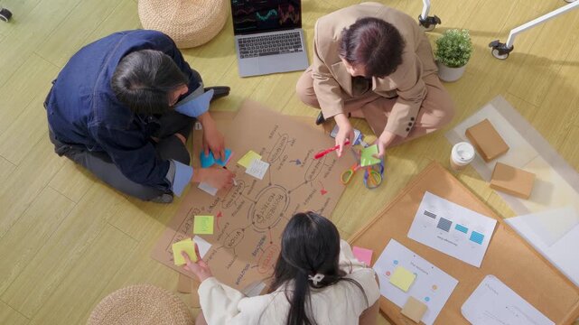 A creative team collaborates casually on the office floor, sketching a business mind map on kraft paper surrounded by colorful sticky notes, charts, and a laptop displaying data analytics.