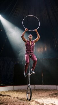 Selbstbewusster m&auml;nnlicher Zirkusartist balanciert auf einem hohen Einrad in der Manege und h&auml;lt einen Ring &uuml;ber dem Kopf