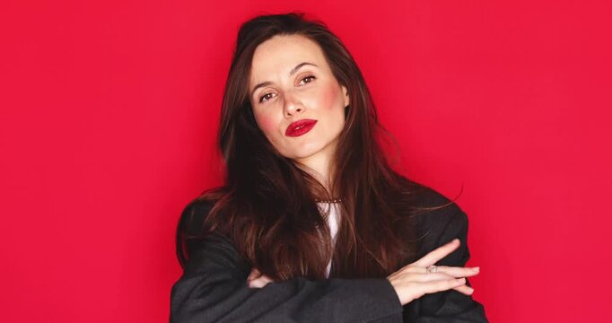 Woman with long dark hair and vibrant red lipstick fixing her hair and crossing arms looking confidently and seriously with calm facial expression isolated on solid red studio background