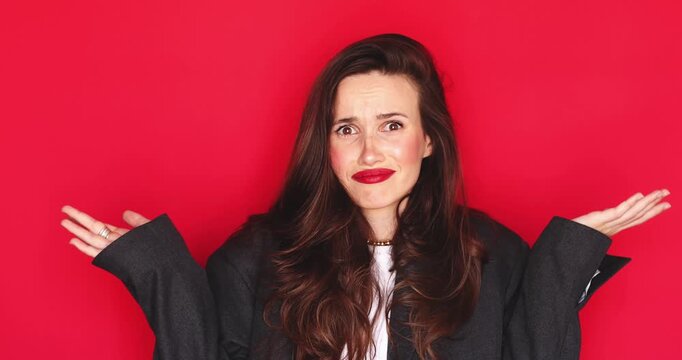 Confused woman with long dark hair and vibrant red lipstick shrugging shoulders feeling doubtful and uncertain with negative grimace expression isolated on a solid red studio background close up video