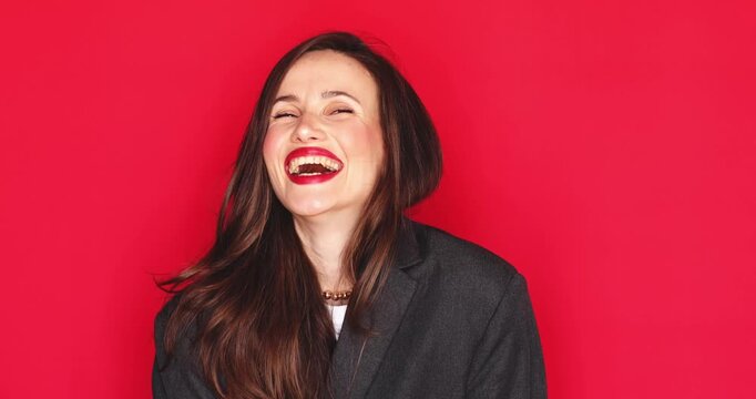 Attractive young woman with long dark hair and vibrant red lipstick laughing out loud with a happy and joyful facial expression crossing arms isolated on solid red studio background close up portrait