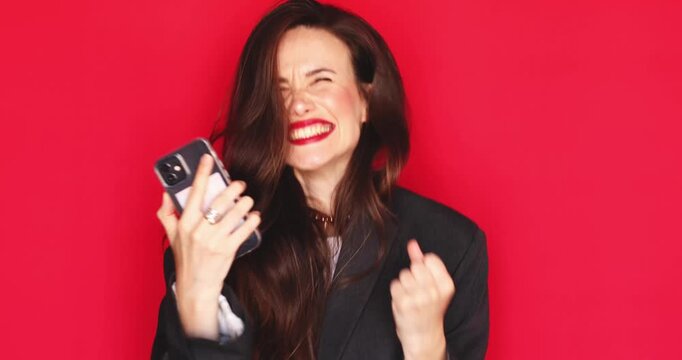 Excited young woman with long dark hair and vibrant red lipstick holding smartphone reacting with thrilled surprise wide open mouth happy joyful expression laughing winning celebrating good news