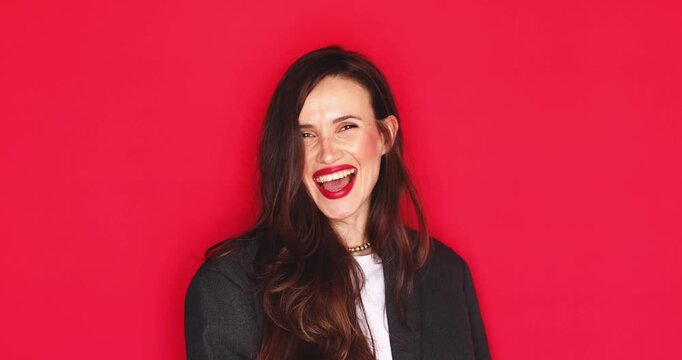 Attractive young woman with long dark hair and vibrant red lipstick laughing out loud with a happy and joyful facial expression showing teeth isolated on a solid red studio background close up 