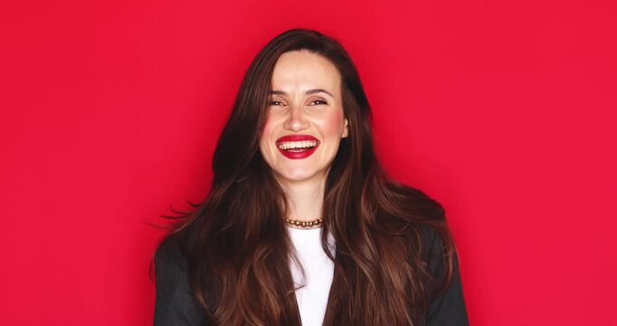 Attractive young woman with long dark hair and vibrant red lipstick showing mini finger heart gesture smiling broadly with a happy and joyful facial expression isolated on solid red studio background 