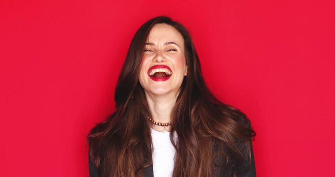 Attractive young woman with long dark hair and vibrant red lipstick showing intense surprise with open mouth and celebrating success with clenched fists and joyous laughter isolated on red background
