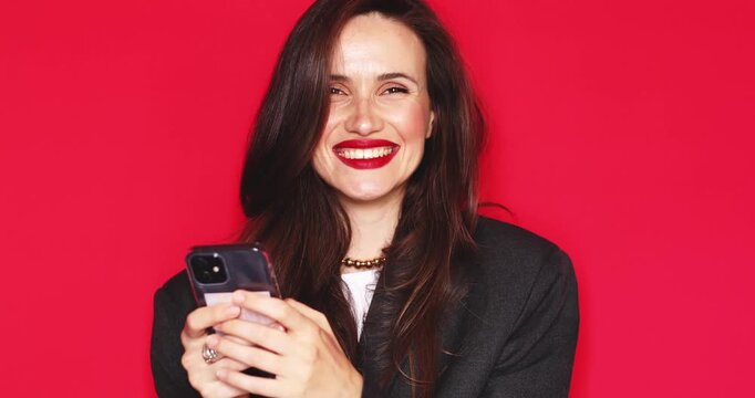 Attractive young woman with long dark hair and vibrant red lipstick looking at smartphone with happy joyful smiling facial expression laughing cheerfully isolated on a solid red studio background 