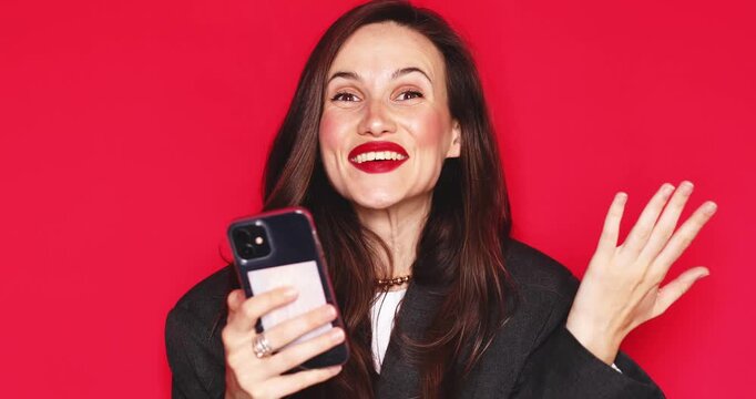 Attractive young woman with long dark hair and vibrant red lipstick holding smartphone receiving great news reacting with extreme surprise open mouth and joyful celebrating laughter isolated on red