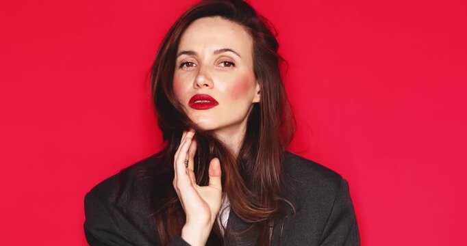 Attractive young woman with long dark hair and vibrant red lipstick looking at camera fixing hair with a thoughtful and calm facial expression isolated on a solid red studio background close up 