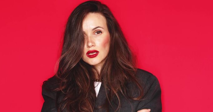 Attractive young woman with long dark hair and vibrant red lipstick looking at camera fixing hair with a thoughtful and calm facial expression isolated on solid red studio background close up portrait