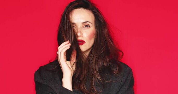 Attractive young woman with long dark hair and vibrant red lipstick looking thoughtfully at camera fixing hair with a calm and serious facial expression isolated on a solid red studio background 