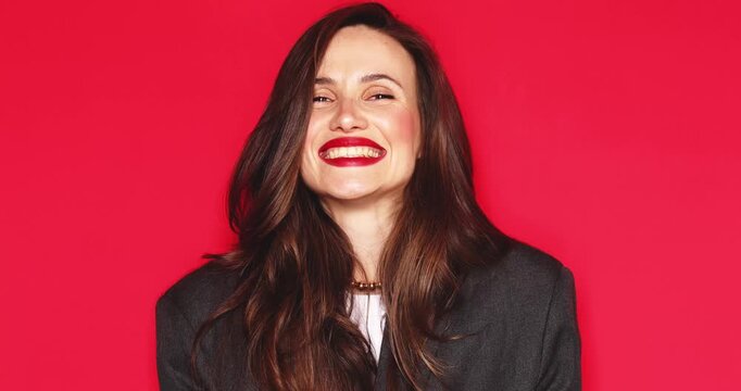 Attractive woman with long dark hair and vibrant red lipstick pointing fingers at camera with playful expression and friendly joyful smiling facial expression isolated on a solid red studio background