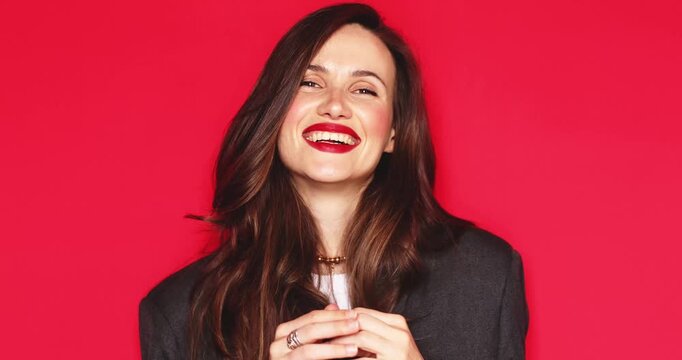 Attractive young woman with long dark hair and vibrant red lipstick looking at camera and smiling broadly with happy joyful facial expression while hiding behind hair on a solid red studio background 